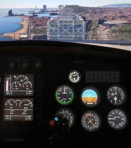Cockpit of small airplane; instrument panel visible as coastline and cityscape with tall white building seen ahead beyond the runway.
